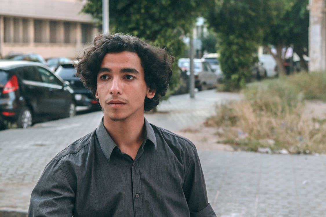 the image is a medium closeup portrait of a young man with curly brown hair. he is wearing a dark gray buttondown shirt. he appears to be of middle eastern or arab descent.  he's standing outdoors on a city street, with several parked cars and buildings visible in a softly blurred background. his expression is somewhat pensive or thoughtful, and he is looking off to the side, away from the camera. the overall mood is relaxed yet slightly introspective.  the shallow depth of field keeps the focus sharply on the young man while softening the background details.