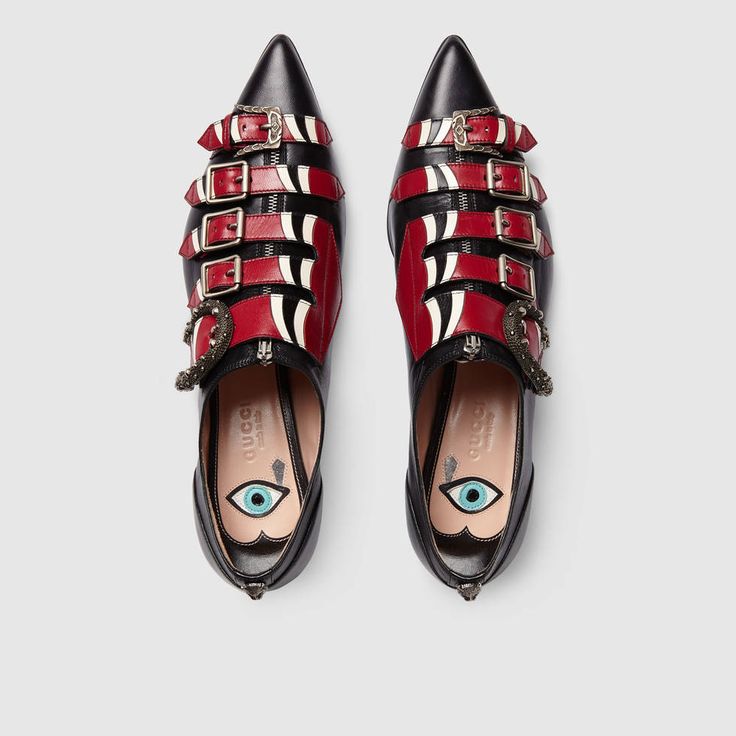 verhead shot of a pair of gucci women's pointedtoe flats.  the shoes are black and red leather with a snakelike pattern and multiple buckles. they feature a zipper closure and a whimsical evil eye detail on the insole.  the shoes appear to be made of highquality leather and are clearly designer footwear.