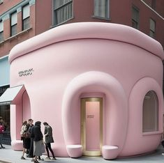 a bright pink, uniquely shaped building stands on a city street. it appears to be a retail store, with its entrance marked by goldtrimmed double doors and a small group of people gathered outside. the building's unusual, rounded design gives it a playful and modern aesthetic.