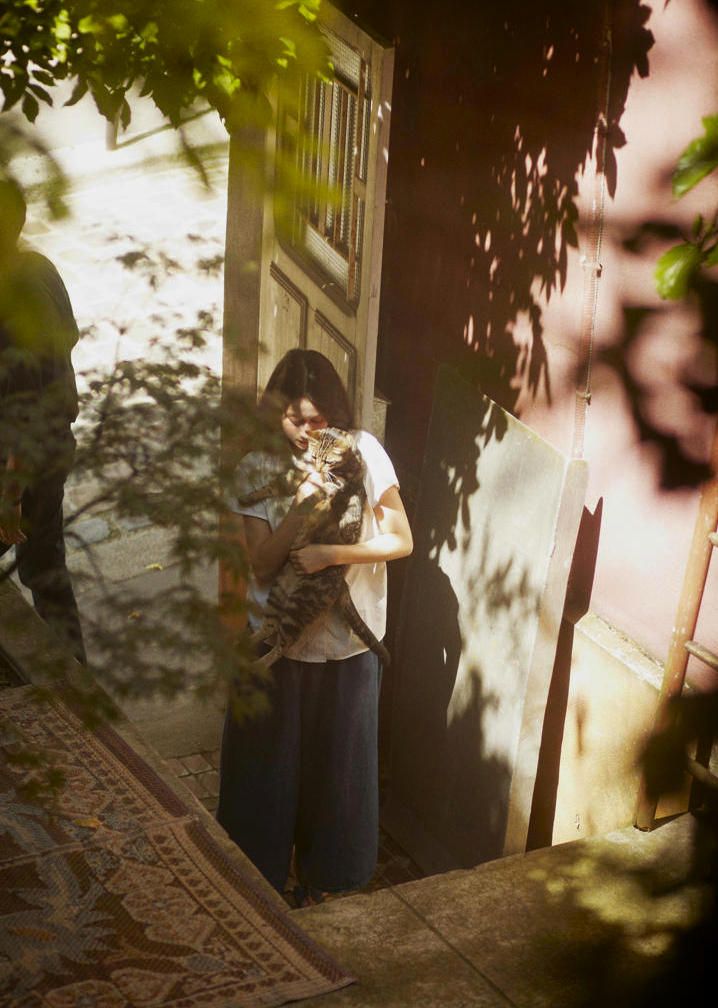 a woman is gently holding and embracing a tabby cat in a sunlit doorway.  she is standing on steps, and the background shows a building exterior with leafy trees casting dappled shadows. the woman is wearing loose, wideleg pants and a lightcolored top.  a patterned rug is partially visible on the steps. the overall atmosphere is calm and peaceful.
