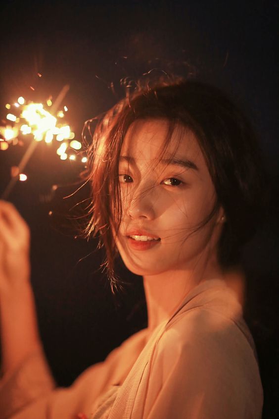 a woman, brightly lit by a sparkler she holds in her right hand, smiles at the camera. the woman has short dark hair and is wearing a peachcolored top. the background is dark.