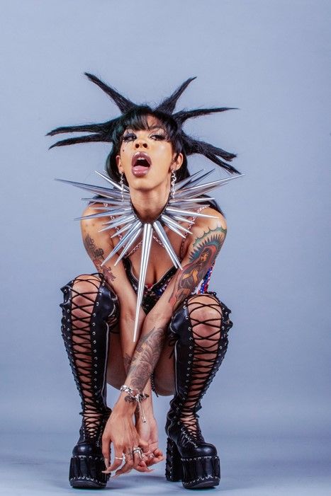 a woman with a punk rock style is sitting with her legs crossed, her back straight, and her face tilted upward. her hair is styled into sharp points, and she is wearing a silver spike necklace. she has tattoos on her arms and is wearing black laceup boots with a thick platform. she is wearing minimal makeup, with bold eyes and dark lipstick. she has a confident and edgy look.