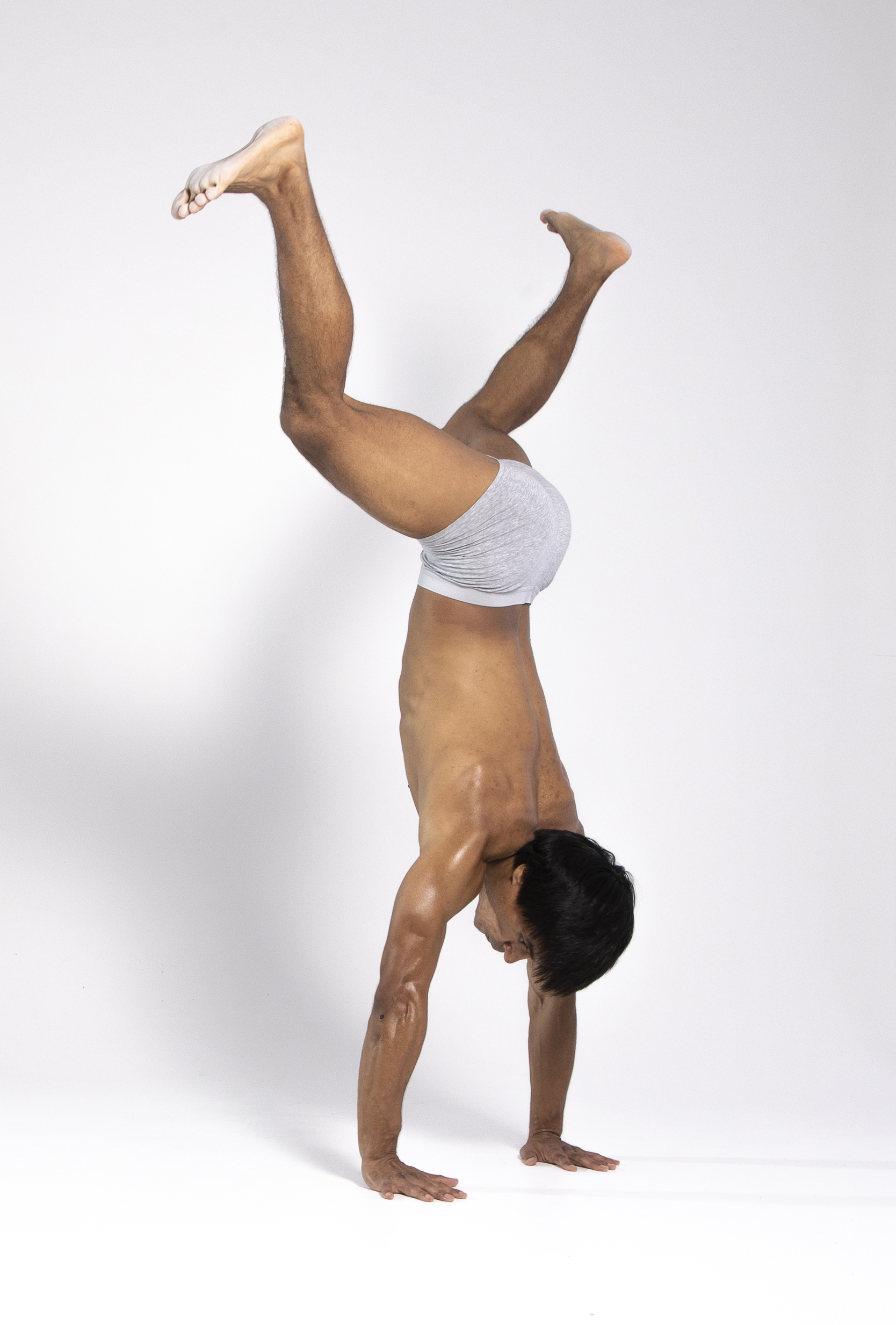 a tanned man wearing grey underwear is performing a handstand in a studio setting. his body is positioned vertically, with his hands flat on the white floor and his legs angled upwards in a split position. the man's head is pointed downwards, and his hair is dark. his body appears to be welltoned, and he is sweating. the background is plain white, creating a clean and focused image.