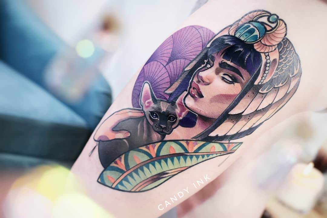a neotraditional style tattoo on someone's upper arm. the tattoo depicts a woman with dark hair and an elaborate egyptianinspired headdress holding a hairless sphynx cat.  the woman and cat are set against a backdrop of geometric patterns and a purple fanlike shape. the tattoo artist's signature "candy ink" is visible in stylized font at the bottom right.