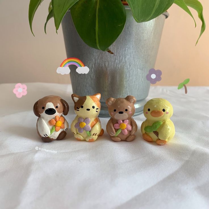 the image shows four handmade clay animal figurines lined up on a white surface with a potted plant in the background. from left to right, there is a brown and white dog holding an orange flower, a calico cat holding a purple flower, a brown bear holding a pink flower, and a yellow duck holding a green leaf. there are also digital graphics of a rainbow, flowers, and a leaf floating in the air.