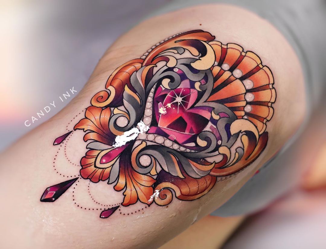 a closeup image of a freshly done tattoo on the back of someone's leg, just above the knee.  the tattoo is an ornate, stylized heartshaped jewel with gold and white scrollwork and three dangling jeweled charms. the tattoo has a realistic, 3d appearance due to the shading and color work. the colors are vibrant and the lines are clean. the tattoo artist's watermark "candy ink" is visible in the upper left corner of the image.