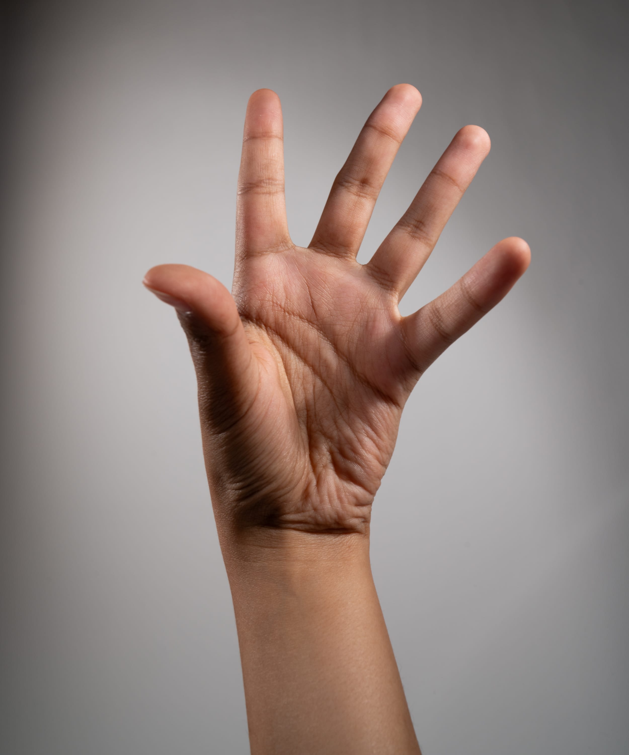 a closeup image of a raised hand against a gray background, illuminated by studio lighting. the hand is extended with fingers spread, showcasing the palm and intricate lines of the skin.