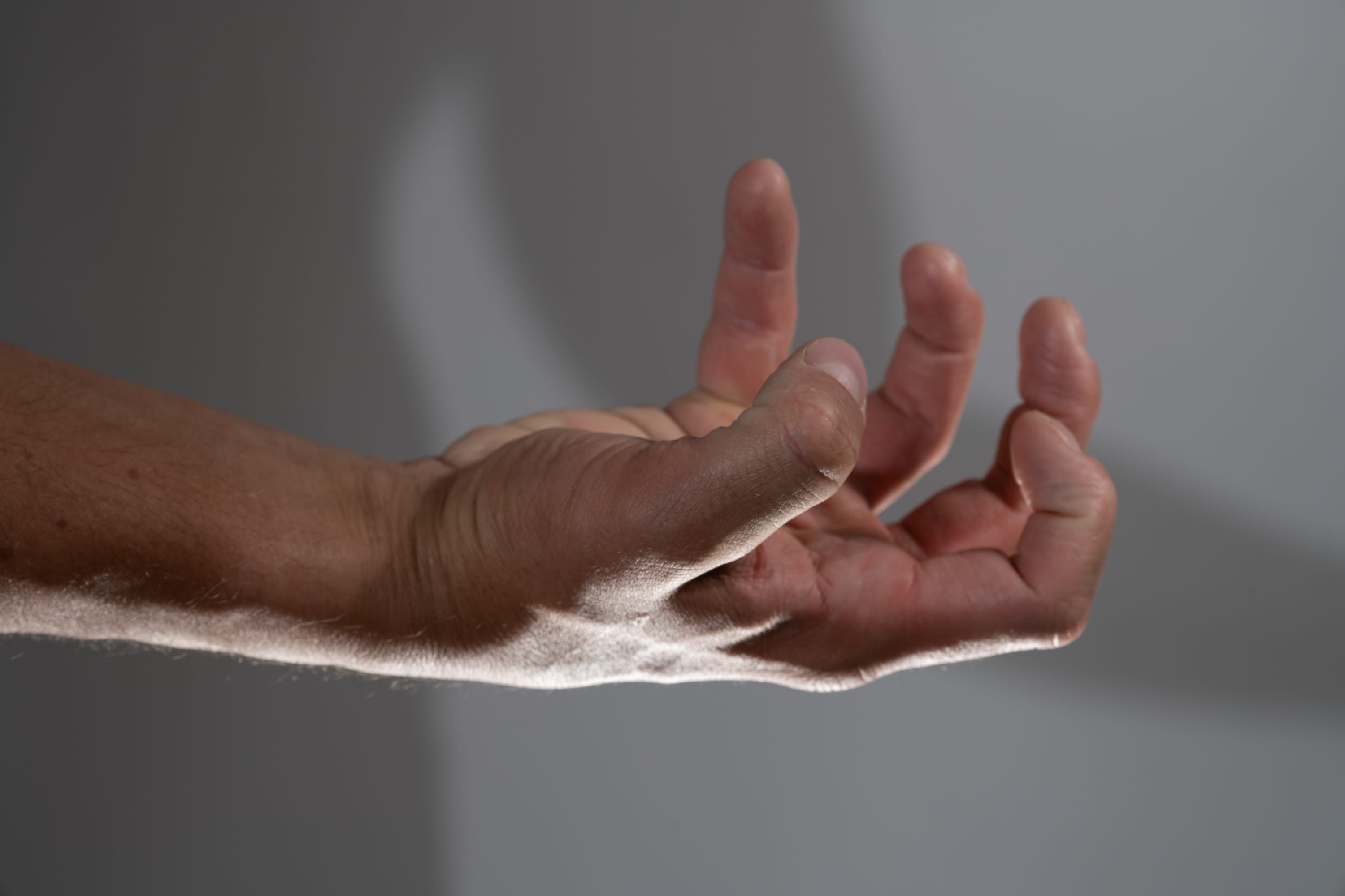 a closeup side view of a man's outstretched hand with slightly curled fingers against a gray background. the hand is lit by a soft light source, creating subtle shadows that accentuate the contours of the skin and fingers.