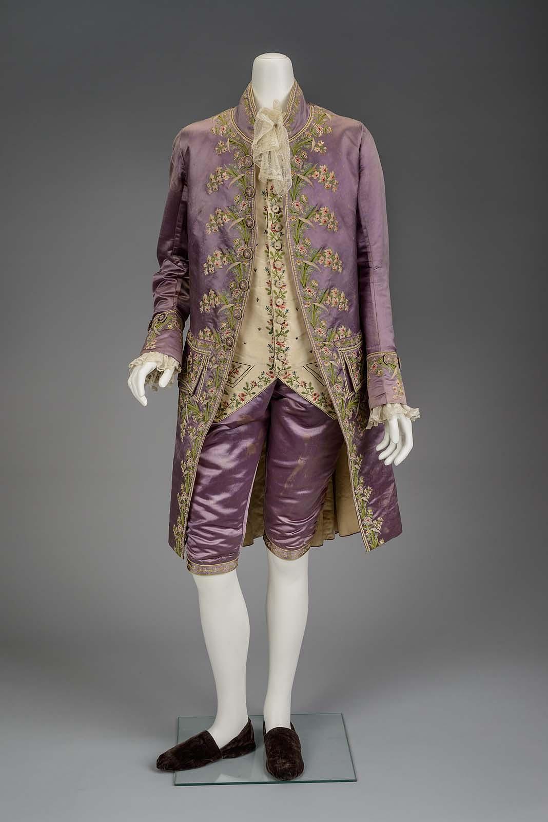 a mannequin displays a formal outfit consisting of a long coat, waistcoat, and kneelength trousers, all adorned with floral embroidery. the coat and trousers are made from a lavendercolored fabric, while the waistcoat is offwhite. the mannequin wears brown slipon shoes. the background is a muted gray.