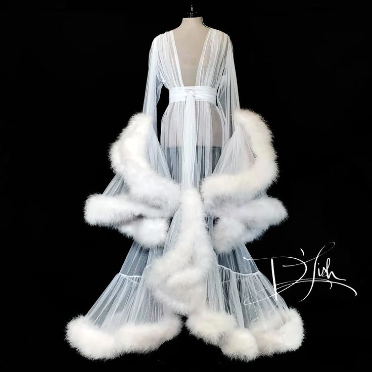 the image shows a luxurious white robe displayed on a mannequin against a black background. the robe is made of a sheer tulle material with a deep vneck and long sleeves. it is adorned with feather trim along the sleeves and hem. a white sash is tied around the waist. the overall style is glamorous and elegant, suggesting it might be used for a bridal boudoir photoshoot or lingerie collection.