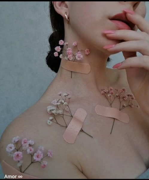 closeup image of a woman's shoulders and neck adorned with small pink flowers and light pink bandaids. the flowers are delicately placed, creating a soft and feminine aesthetic. the woman's nails are painted with a soft pink nail polish, complementing the overall pastel color scheme. she is wearing gold hoop earrings. the overall vibe is delicate, minimalist and elegant.