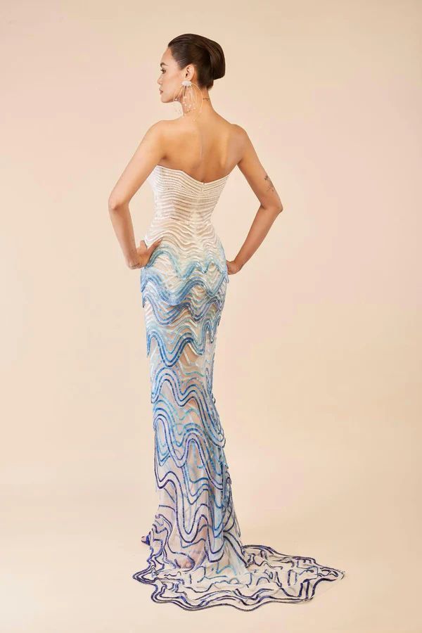 a woman is standing with her back to the camera, wearing a strapless white and blue gown with a wavy pattern. the gown has a train. she has an updo hairstyle and is wearing earrings. she has her hands on her hips. the background is a light peach color.