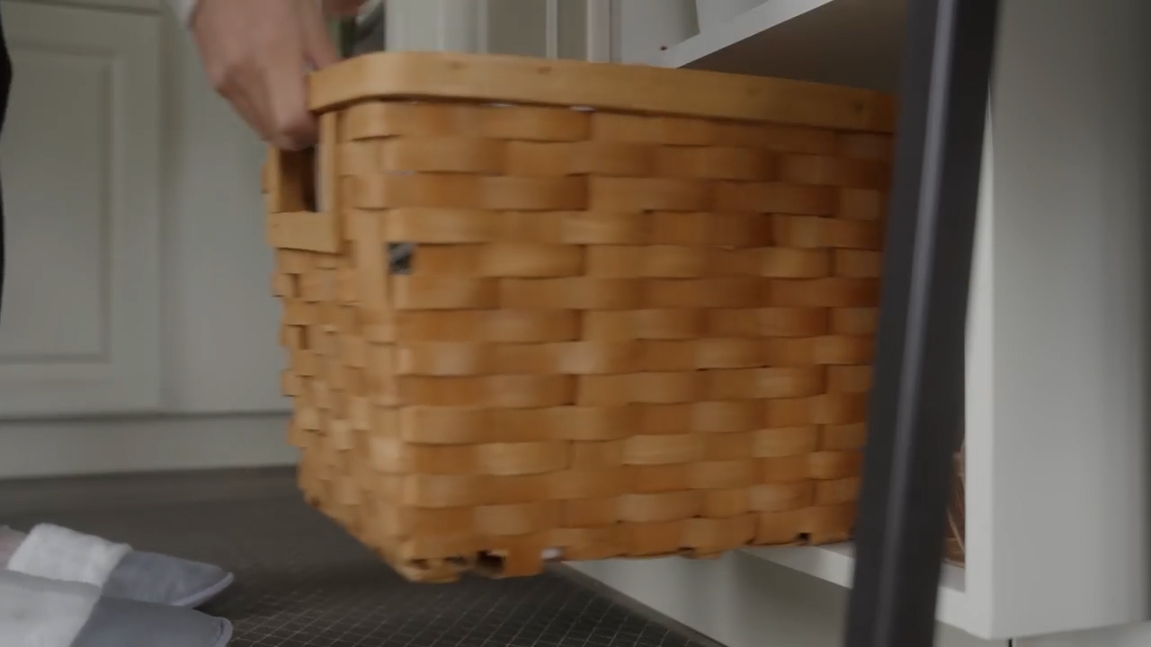 a person is shown pulling a woven basket out from a shelf.  the basket and shelf are light wood tone.  the shelf is supported with a matte black metal frame.  the person is wearing gray slippers and black pants.  the floor is covered with large format gray tiles.  the walls are white.