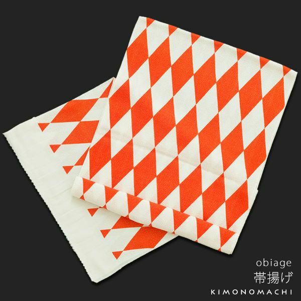 the image shows a folded obiage, a japanese kimono accessory, with an orange and white diamond pattern. it is associated with the kimonomachi brand and has japanese text that reads "obiage" and "帯揚げ" (obiage). the fabric is folded and lies on a black background.