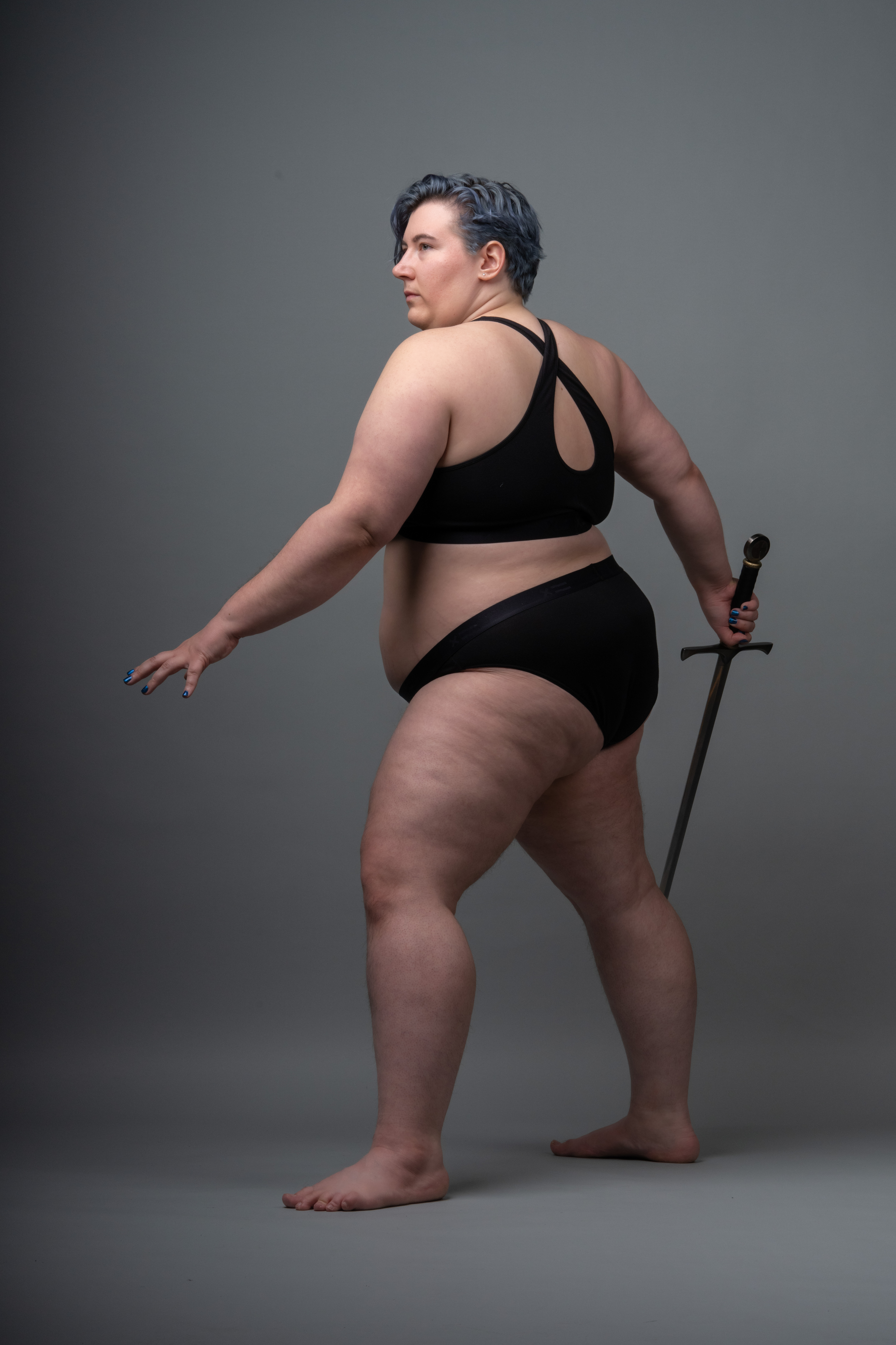 a plussize woman with short, blue hair is pictured in a studio setting. she's wearing a black sports bra and black underwear and standing barefoot on a grey background. she's holding a sword by her side and has a serious expression while looking over her shoulder. her other arm is extended as if in a defensive posture. the photo captures her entire body in a side view.