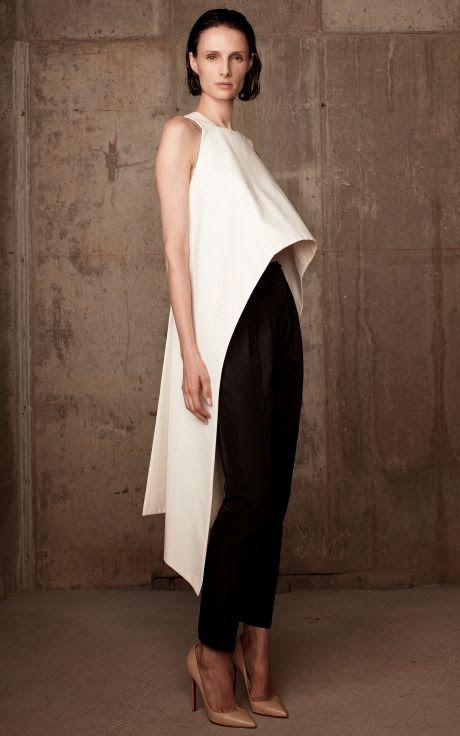 a woman in a minimalist outfit is shown in a studio setting. she wears a white asymmetrical dress over black slimfit pants, paired with nude high heels. the background is a textured concrete wall.