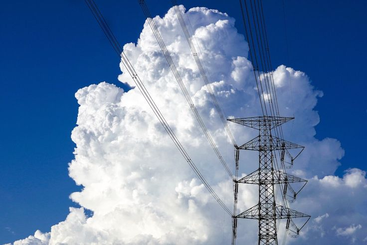 a power transmission tower stands against a backdrop of puffy white cumulus clouds in a bright blue sky. power lines extend from the tower, intersecting the clouds.