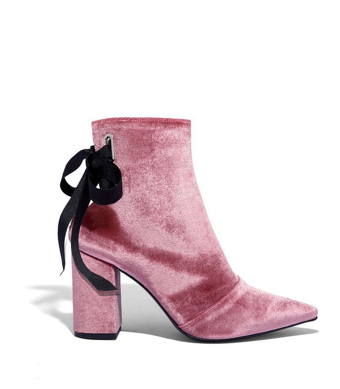a single pink velvet ankle boot with a black ribbon tied at the back is presented against a white background. the boot has a pointed toe and a block heel. the velvet texture is evident, and the black ribbon adds a decorative element to the design.
