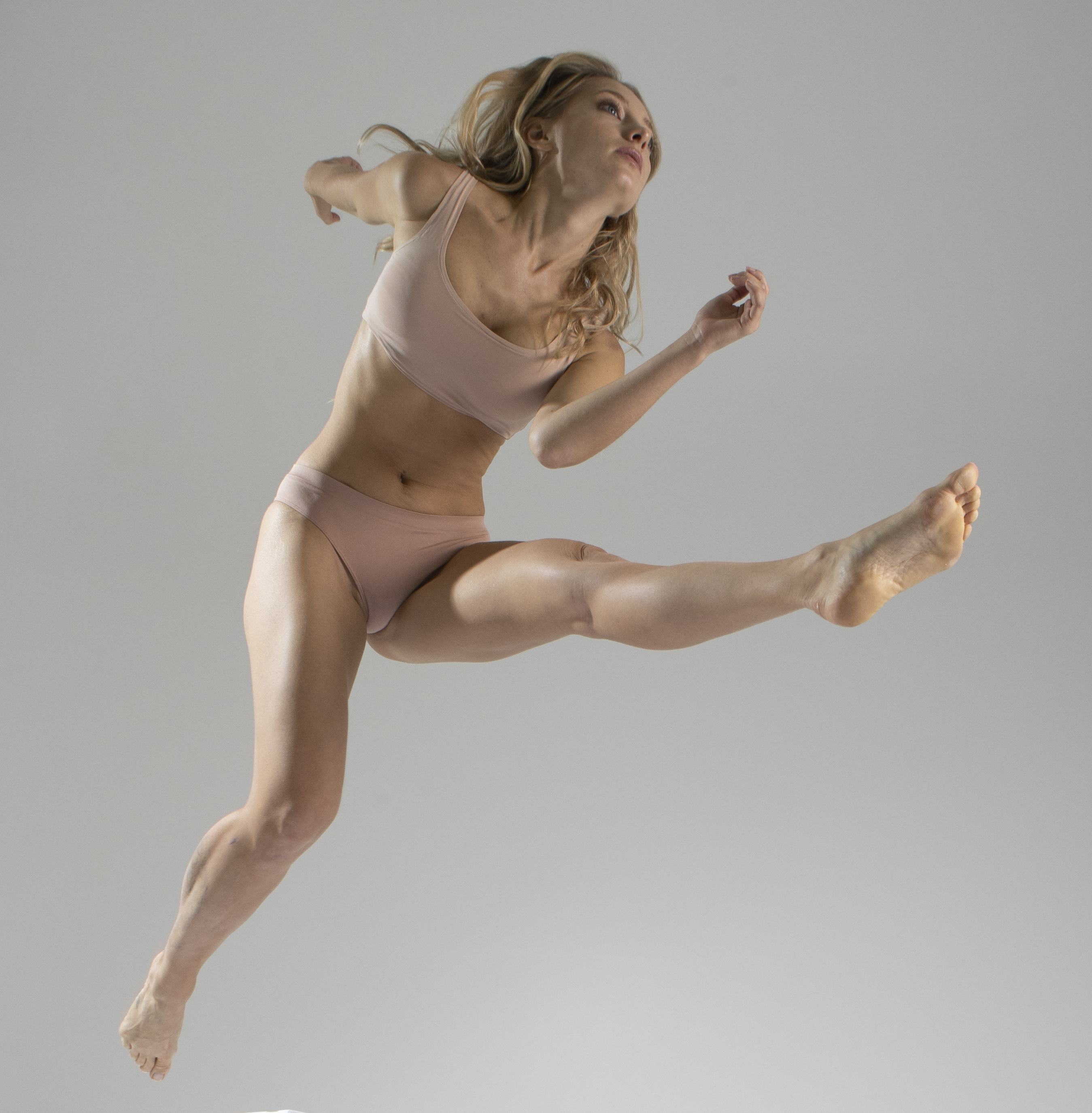 a woman wearing a nudecolored bra and underwear is captured in a midair jump. she is barefoot and her legs are outstretched in a split position, with one leg extended forward and the other backward. her body is angled slightly upwards, and her arms are extended for balance. she has blonde hair and a determined expression on her face. the background is a light grey, suggesting a studio setting. the image conveys energy, athleticism, and body awareness.