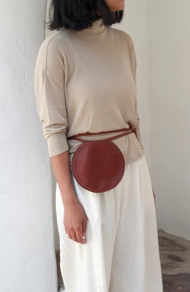 a woman is standing with her body turned slightly to the right, wearing a beige turtleneck sweater and white, loosefitting pants. a round, brown leather purse is attached to a thin leather belt around her waist, positioned over her left hip. the background includes a white wall and a glimpse of a floor.