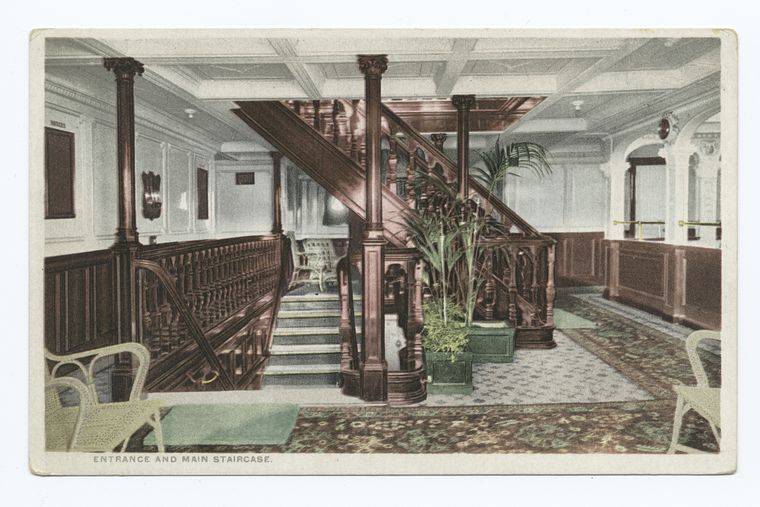 this is a postcard of the entrance and main staircase, presumably of a ship. the staircase is made of wood and has a handrail and pillars. there are plants in pots at the base of the staircase. there is furniture in the form of chairs. the floor is covered in carpeting.