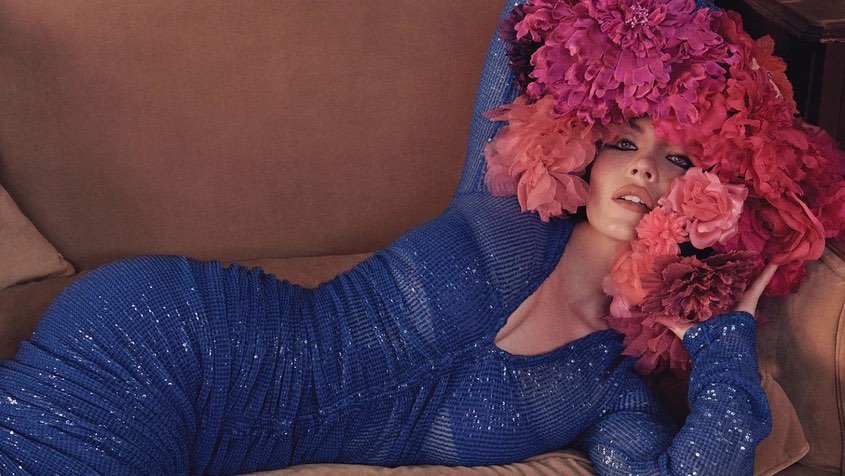 a glamorous woman, possibly a model, is lying down on a brown couch.  she is wearing a stunning, formfitting, sequined blue dress with long sleeves and a vneck.  her hair is obscured by a large, dramatic headpiece crafted from vibrant pink and red flowers, predominantly peonies.  the lighting and overall aesthetic are soft and luxurious, evoking a highfashion editorial style. the woman's makeup is flawless, enhancing her features. the image conveys a sense of elegance, sensuality, and sophistication.