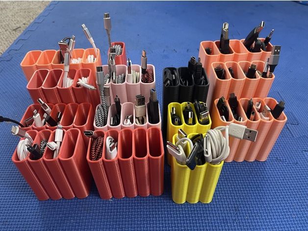 the image shows several 3dprinted cable organizers, each with multiple compartments to hold various types of charging and data cables.  the organizers are stacked together and are in shades of pink, black, and yellow.  the cables are neatly arranged and organized within each compartment. the organizers appear to be designed for desktop or other workspaces to keep cords from becoming tangled. the background is a blue mat.