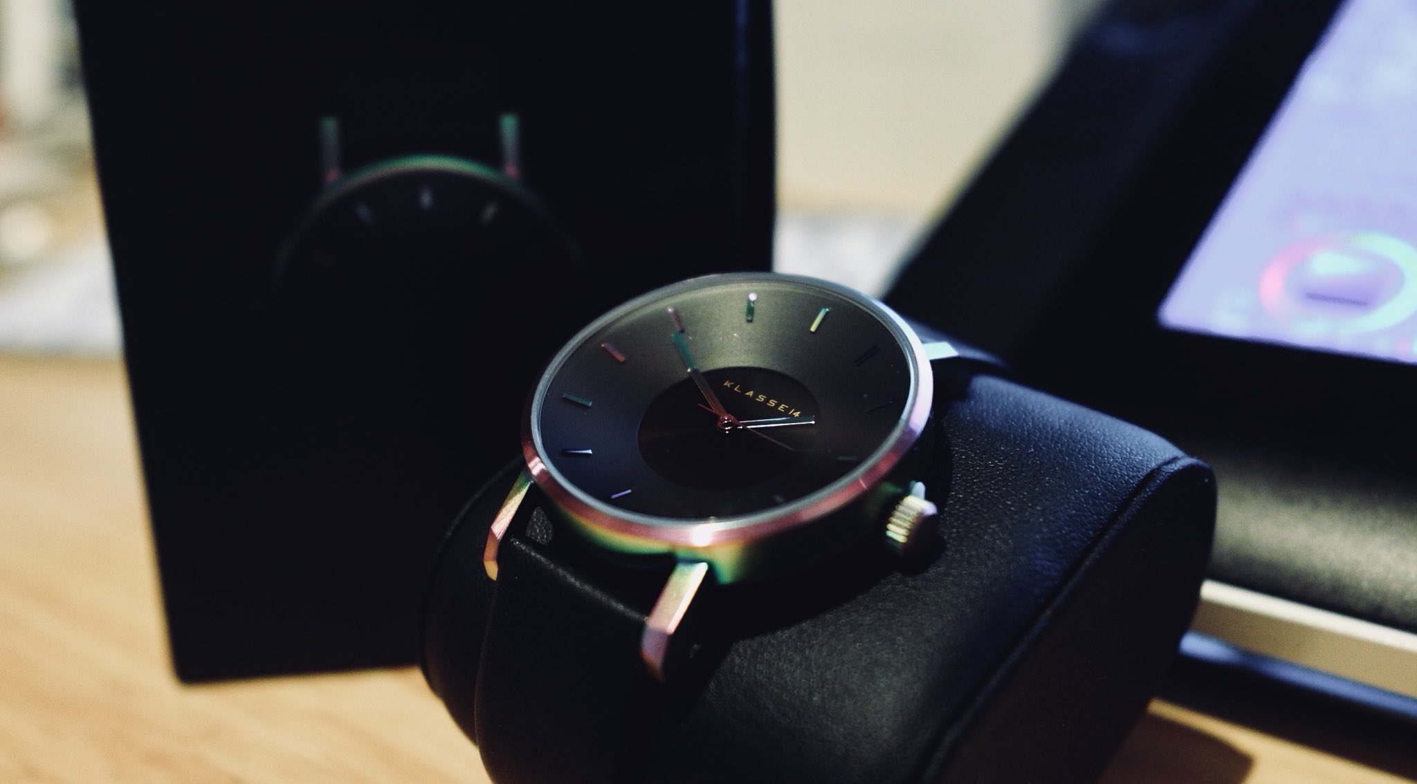 this is a closeup shot of a sleek and modern wristwatch displayed on a black watch cushion. the watch features a minimalist design with a black face, silver hands, and a black leather strap. the watch is presented in a black watch box with the brand name "klassik" visible on the watch face. the background is slightly out of focus, drawing attention to the watch as the central subject. the lighting is soft and diffused, creating a sense of luxury and sophistication.