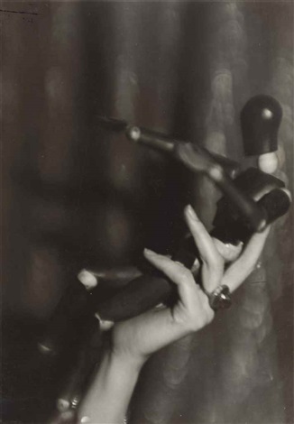 a closeup black and white photograph shows a hand delicately holding a small, stylized mannequin or doll. the mannequin is positioned as if in midaction, suggesting movement and a narrative. the background is blurred and indistinct, focusing attention on the hand and the figure. the overall tone is dark and mysterious, evoking a sense of surrealism or fantasy. the image has a vintage or antique feel, with visible texture and grain.