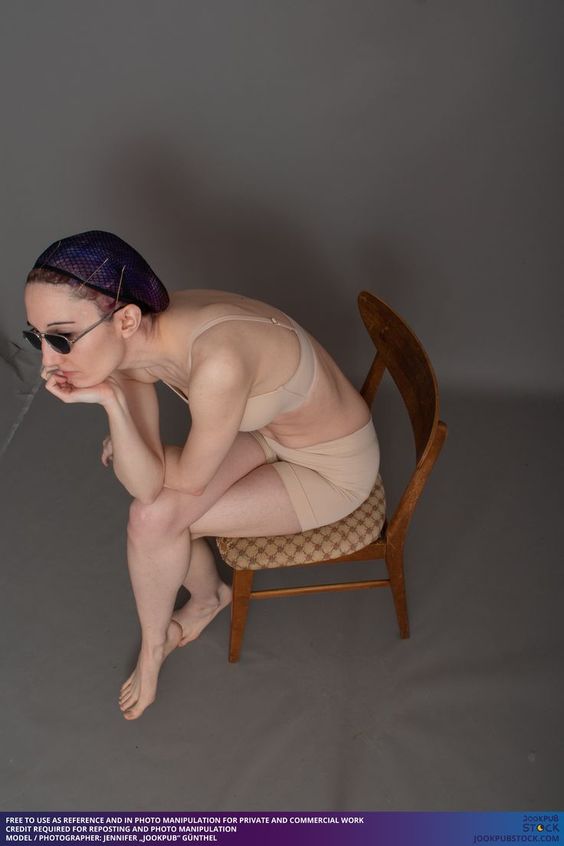 a woman with purple hair wearing sunglasses, a white bra and beige underwear, is sitting on a chair with a floral fabric seat. she rests her head on her hand, lost in thought. she is posing against a gray backdrop, illuminated by artificial lighting.