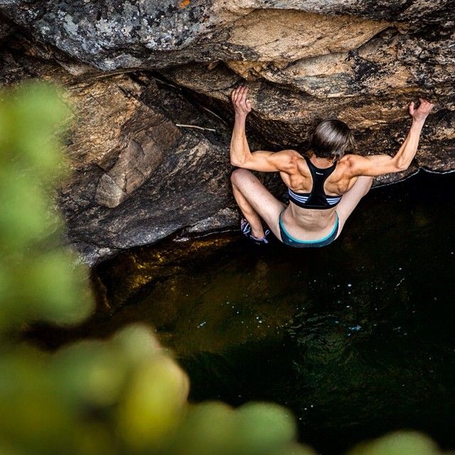 a woman is rock climbing on a rock face that overhangs a body of water. she is positioned with her back to the camera, showcasing her muscular back and arms as she grips the rock. the perspective is from above, looking down at the climber and the water below. the environment suggests a natural setting, with sunlight illuminating the scene.