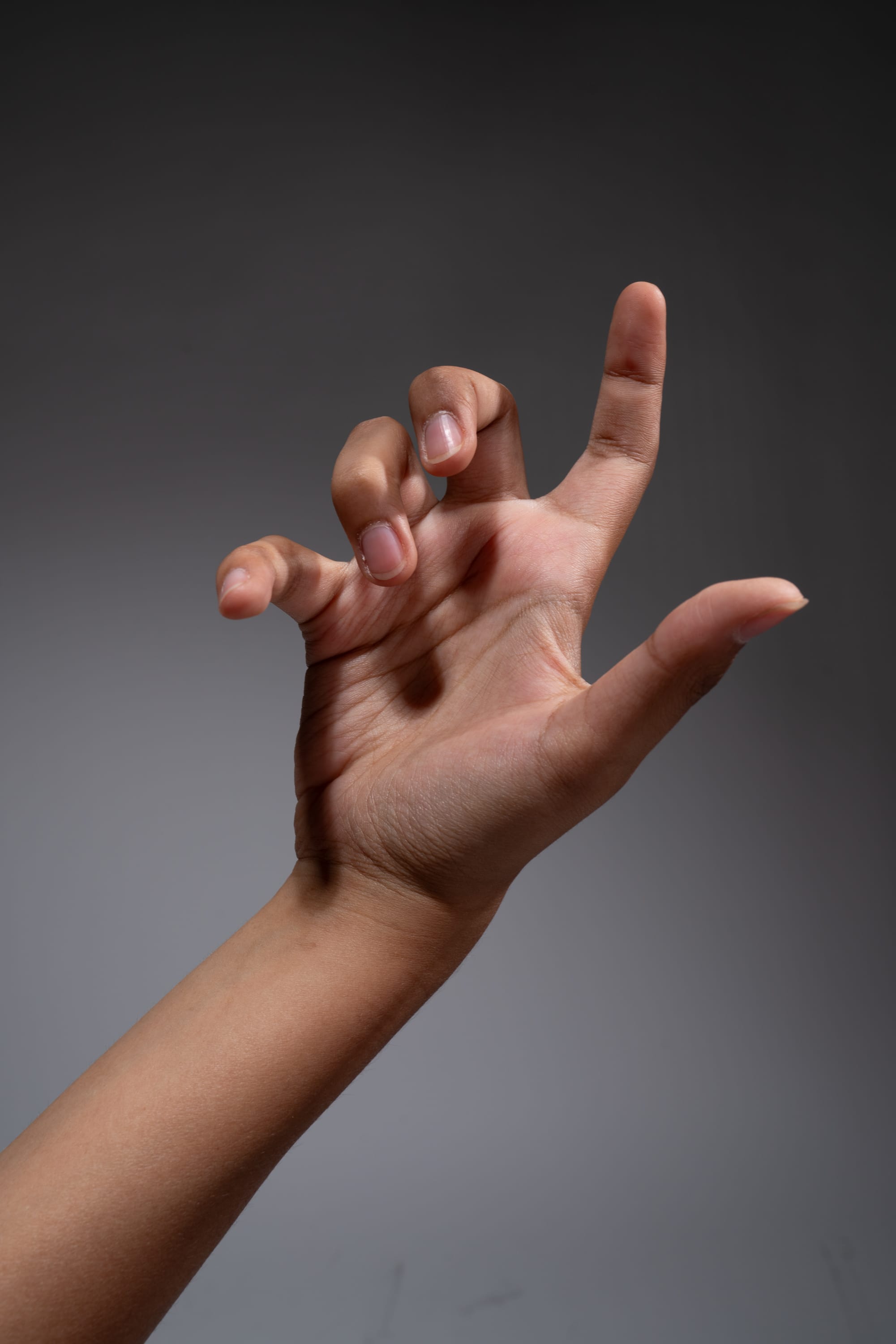 a woman's hand is extended towards the camera, fingers curled in a clawlike gesture against a neutral gray background. the lighting highlights the contours of her hand and arm, emphasizing the intricate details of her skin texture.