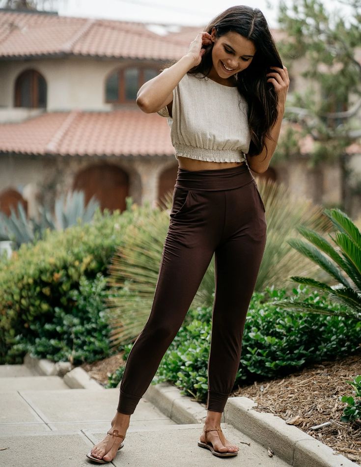 a woman standing on concrete steps in front of a house. she is wearing a beige, cropped, shortsleeved blouse, brown pants, and brown sandals. her hair is long and dark, and she is smiling slightly while looking down. the image appears to have been taken outdoors during the day, as evidenced by the natural lighting.