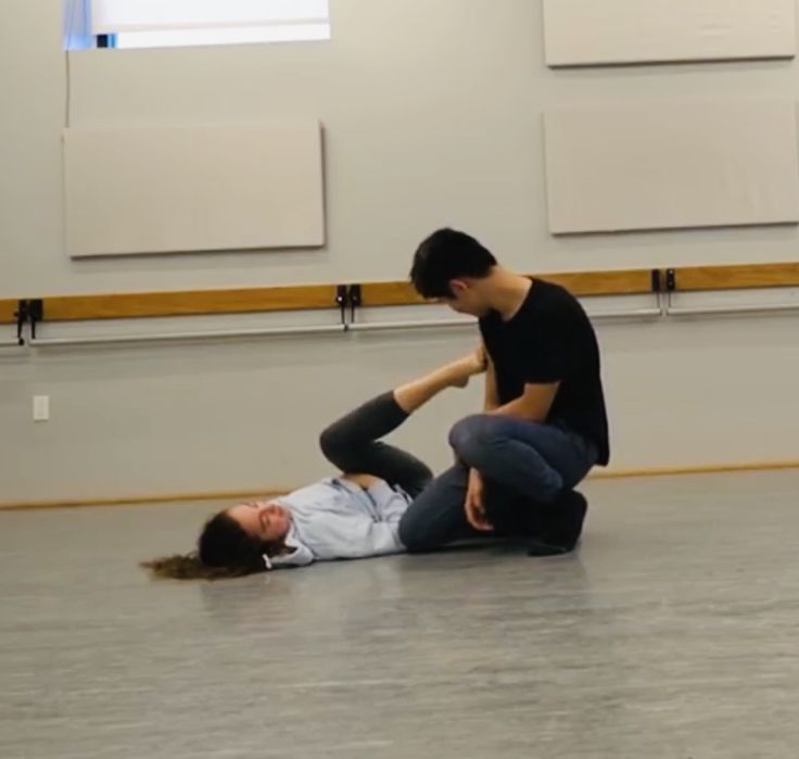 a man and a woman are dancing together in a dance studio. the man is holding the woman's leg as she stretches, and the woman is lying on the floor with her leg extended. they are both wearing casual clothes and are in a modern dance pose. the studio is decorated with a grey floor, and there are a few white panels on the wall. the man has black hair and the woman has brown hair.  they both look focused on their dancing.  there is a ballet bar visible in the background.