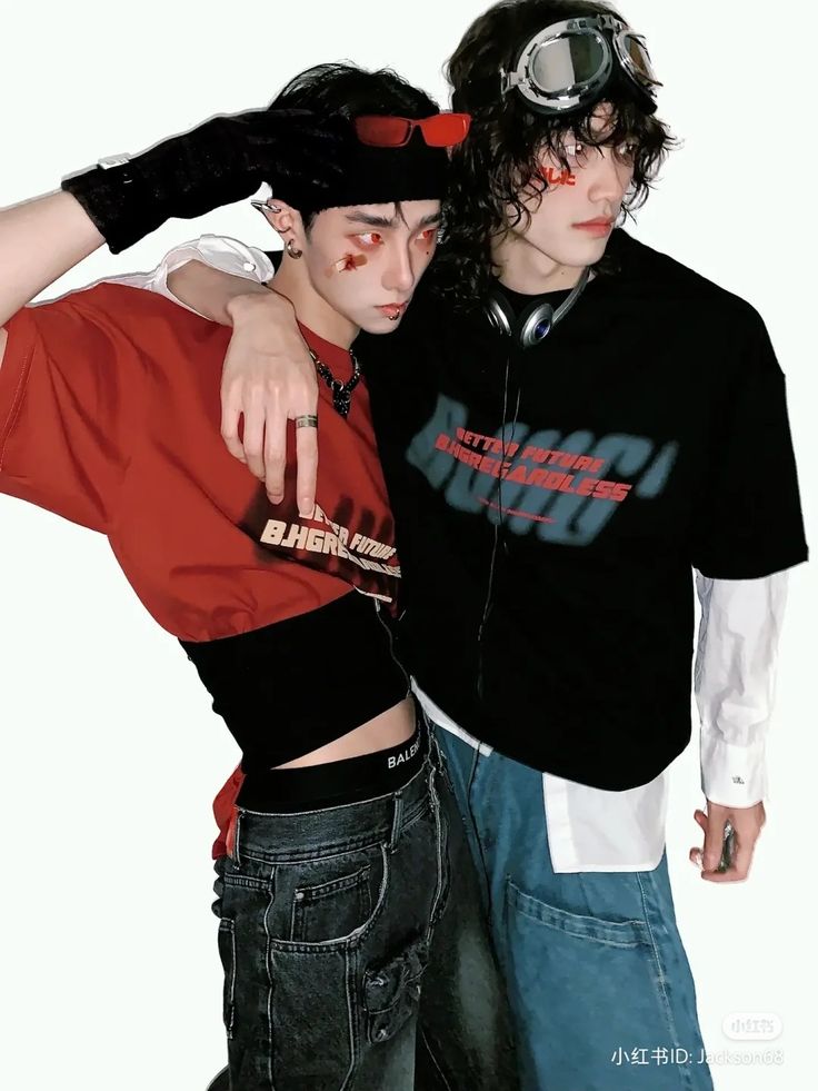 two young men pose against a white background, showcasing an edgy and alternative fashion sense. one wears a red cropped tshirt, revealing a black branded waistband, and black jeans. he accessorizes with redtinted sunglasses and a single black glove. the other wears a black graphic tshirt over a white buttondown, paired with black pants. he sports aviator goggles with a red mark on his face and has dark, curly hair. the studio lighting illuminates the scene, highlighting their distinct styles.