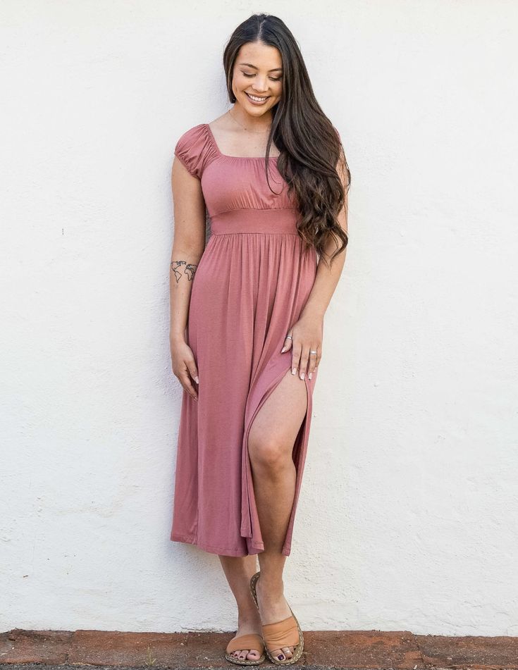 a young woman is standing in front of a white wall, she is wearing a dusty pink midi dress and smiling. the dress has a square neckline, short sleeves, and a slit on one side. the dress is cinched at the waist and flows loosely to her midcalf. she is wearing brown sandals and has long dark hair. the photo is taken from a slightly low angle.
