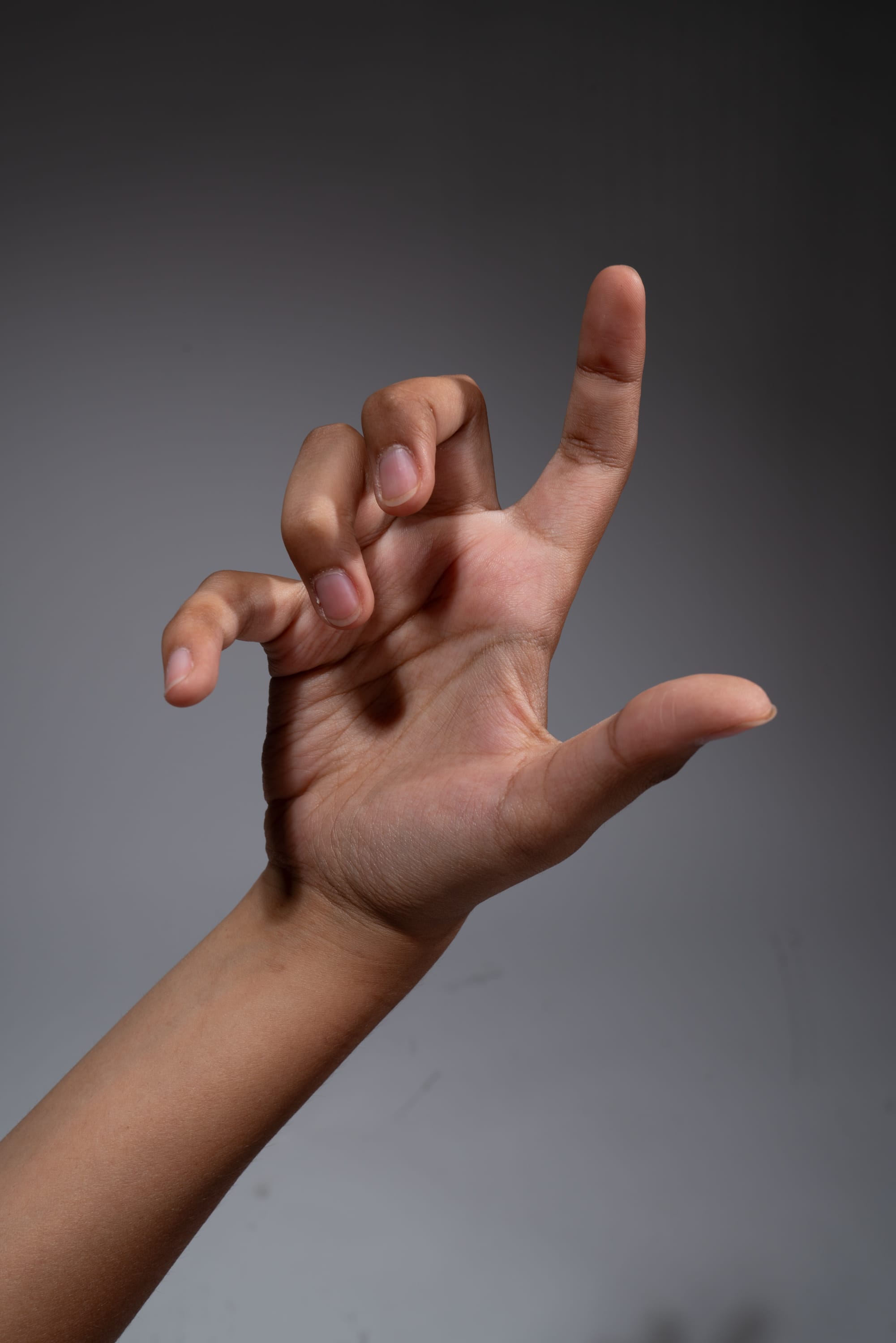 a hand is extended towards the viewer, with the fingers curled in a clawlike gesture. the background is a neutral gray, and the lighting is soft and diffused. the skin tone of the hand is brown.