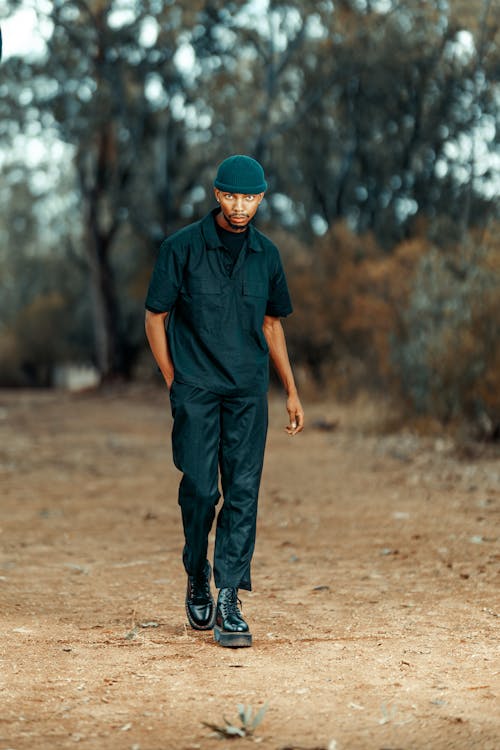 a young man in a dark green hat, shirt, and pants walks along a dirt path in a wooded area. he is wearing black boots and has a serious expression on his face as he looks directly at the camera. the background is blurred but shows the trees and forest floor. the overall feel is one of casual fashion and outdoor lifestyle.