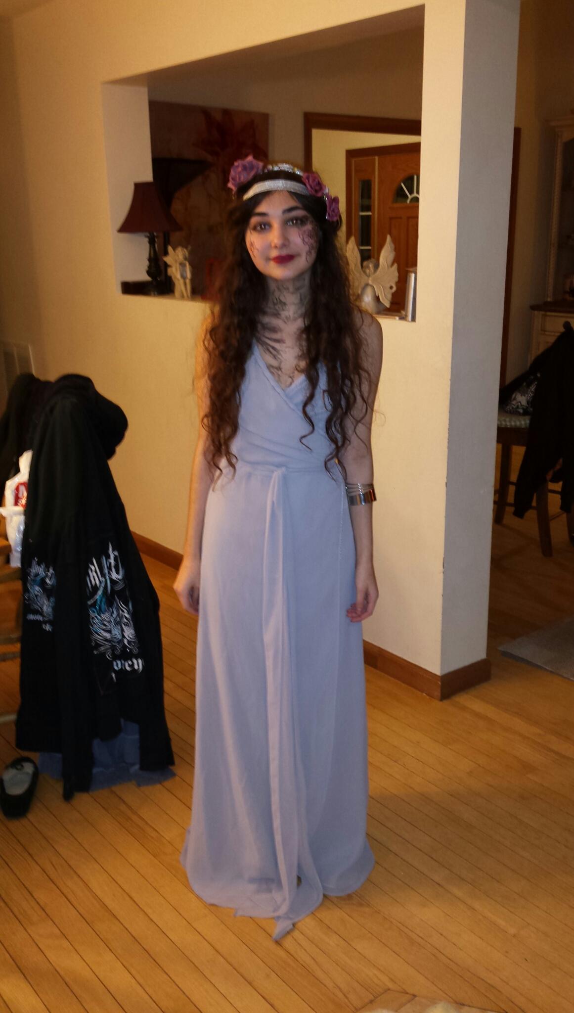 a young woman is standing in a light blue maxi dress. she is wearing a flower crown and has subtle makeup on. she's smiling slightly. the setting appears to be indoors, with a wooden floor. a black hoodie and shoes are on the floor near her feet. there's a doorway and hallway visible in the background, suggesting a home setting. the overall vibe seems like a casual photo, possibly a costume or a casual outfit.