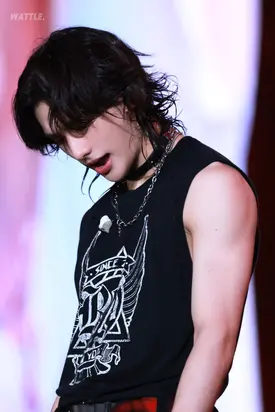 the image is a closeup shot of a young male kpop idol. he has long, dark hair styled in a mullet, and he's wearing a black sleeveless shirt featuring a graphic design and a silver chain necklace. his expression is intense, and his muscular arms are visible. the background is blurred, suggesting a stage performance or similar setting. the image has a "wattle" watermark. the overall aesthetic is edgy and stylish, typical of a kpop performance.