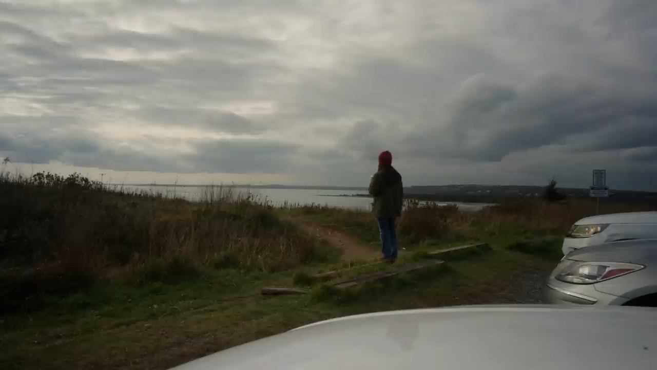 a woman in a red hat stands on a wood plank platform overlooking a grassy cliff to the ocean. there is a dirt pathway leading to where she is standing. it's an overcast day. there is a parking lot with cars to the right. the image is taken from behind a car.