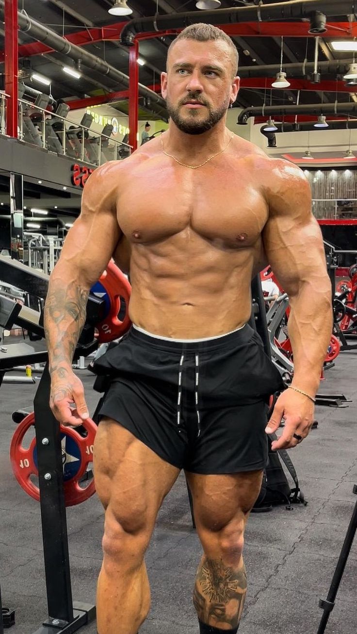 the image shows a muscular male bodybuilder standing in a gym. he's shirtless, displaying a highly developed physique with visible muscle definition in his chest, shoulders, arms, and legs. he's wearing black athletic shorts and has a tattoo on his lower leg. the background shows gym equipment and a partially visible second person in the distance. the overall impression is one of strength, fitness, and athleticism. the lighting and angle are designed to highlight the subject's physique.