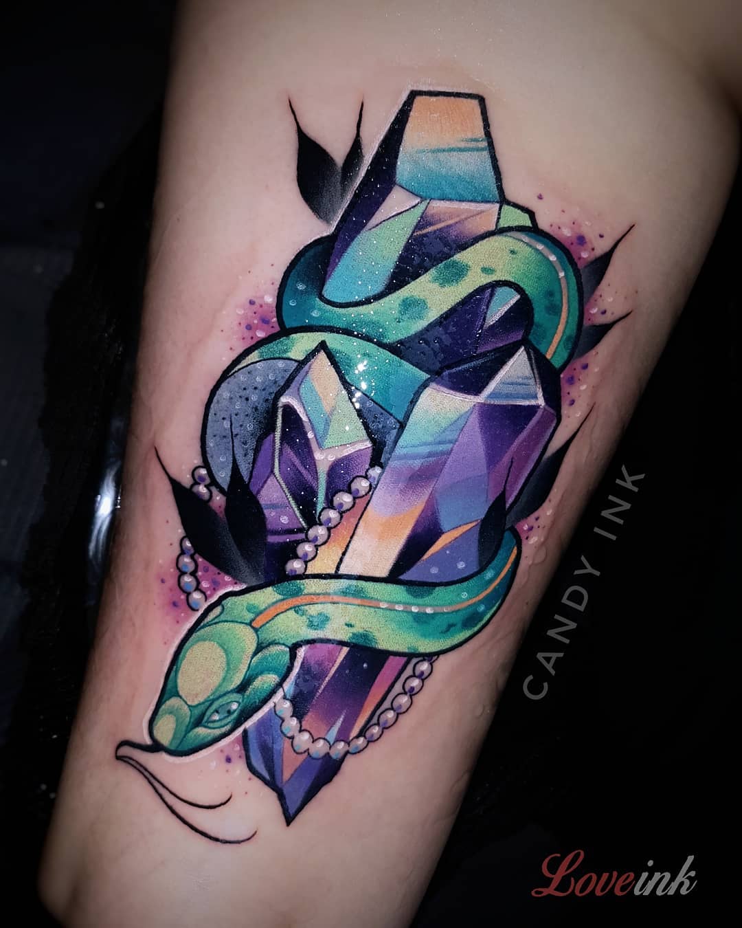 a neotraditional tattoo of a green snake wrapped around some sparkling purple and blue crystals.  there's a string of pearls draped over the design and some dark stylized leaves in the background.  the artist's signature, "candy ink", is written in white cursive on the right side of the image, and the studio's watermark, "love ink", is written in pink cursive script on the bottom right of the image.