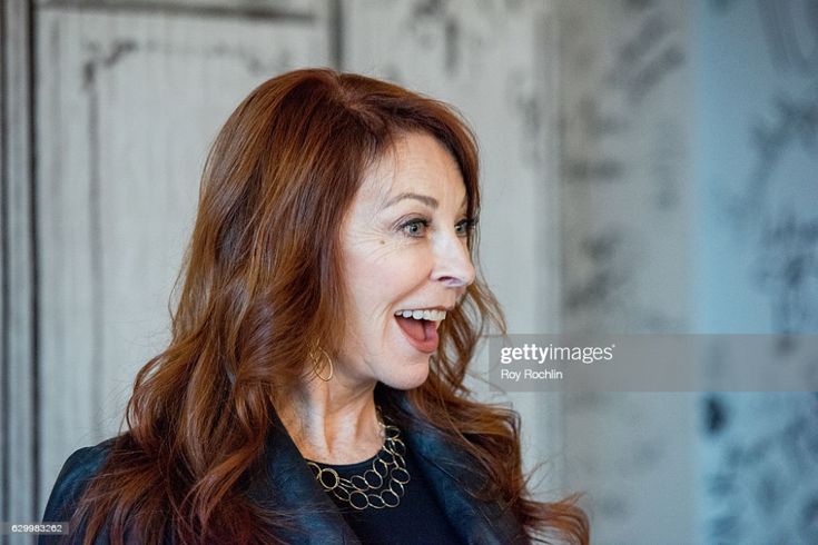 a woman is smiling brightly as she looks to the side in this studio portrait. she has long, curly brown hair, and she's wearing a black jacket and a silver necklace.