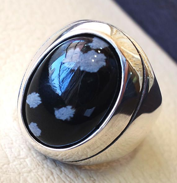 a closeup of a silver men's ring with a black and white oval gemstone. the ring is set in a silver band with a smooth finish. the gemstone is a black stone with white flecks. the ring is laying on a white surface.