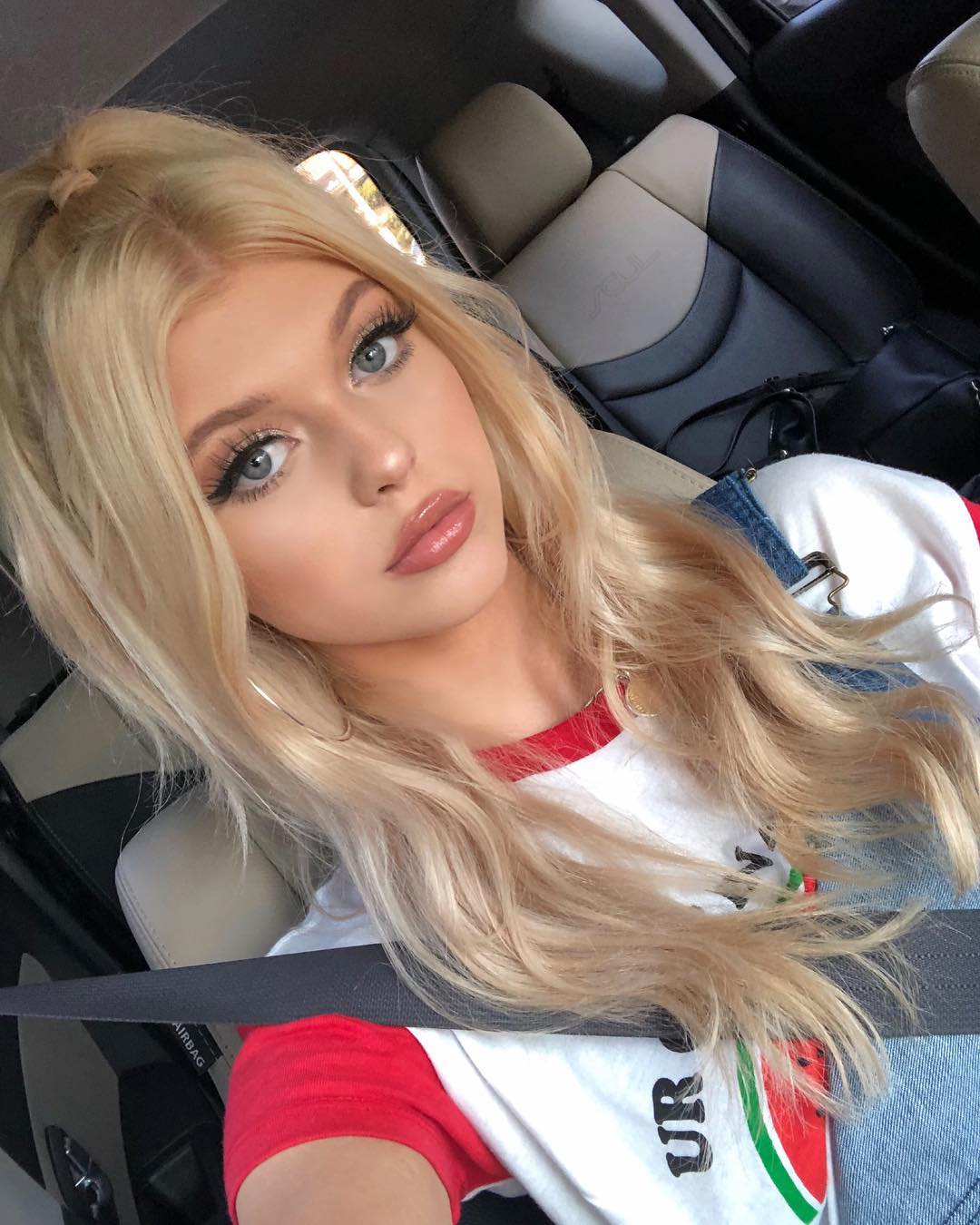 a selfie of a blonde woman wearing a red and white tshirt and a seatbelt in a car. she has her hair styled in a halfup ponytail and is wearing makeup, including eyeliner and lip gloss. the background shows the car's interior, including the seat and door.