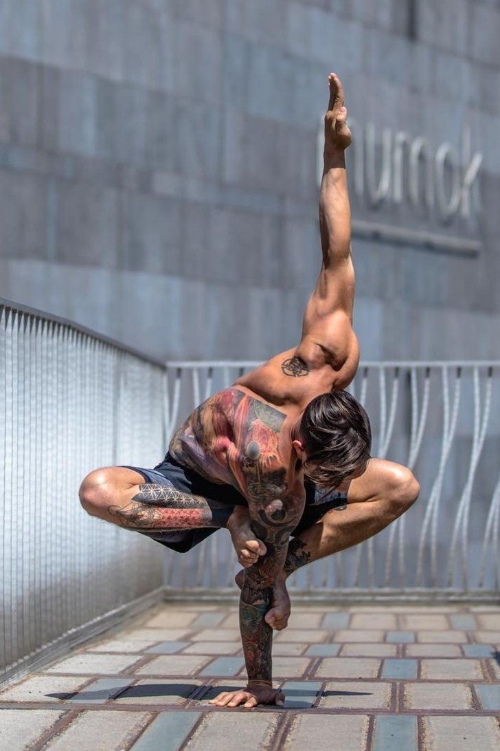 a man is balancing on one arm in a yoga pose. he is tattooed and wearing black shorts. he is standing on a concrete surface in a city. he is reaching up with his other arm.