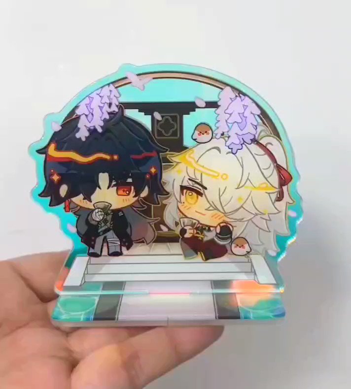 a closeup shot of a chibi anime character acrylic stand, with a male character wearing a black jacket and a female character with white hair and red accents on her outfit, both smiling and looking cute. the background is a colorful abstract pattern and they are standing in a beautiful wooden platform. the stand is being held in a person's hand.