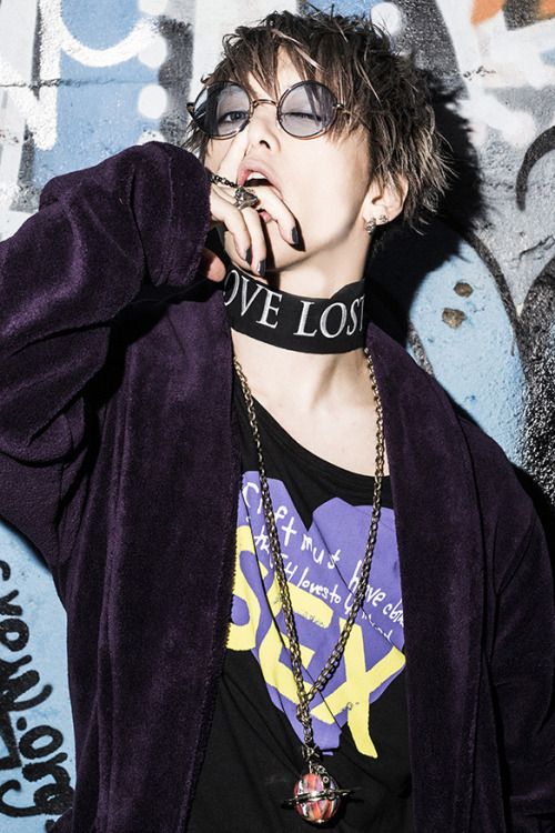a young man is posing in a purple robe. he has a dark choker on that says "love lost" and a gold chain around his neck. he is wearing glasses and has a black tshirt with a large yellow heart on it. his hair is styled with a piece sticking up on top. he is standing in front of a graffiti wall. he has a serious expression on his face and is biting his finger.