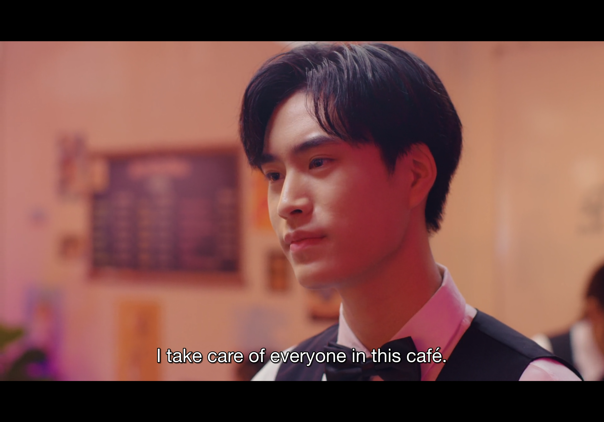 a closeup shot of a young asian man with dark hair and a gentle expression. he is wearing a black vest over a white shirt, suggesting he might be a waiter in a cafe. the background is slightly out of focus, with warm, pinktoned lighting, hinting at a cozy and inviting atmosphere. the text "i take care of everyone in this cafe" is overlaid on the image, indicating that he is a caring and responsible individual, likely speaking these words as a promise or statement about his work ethic.